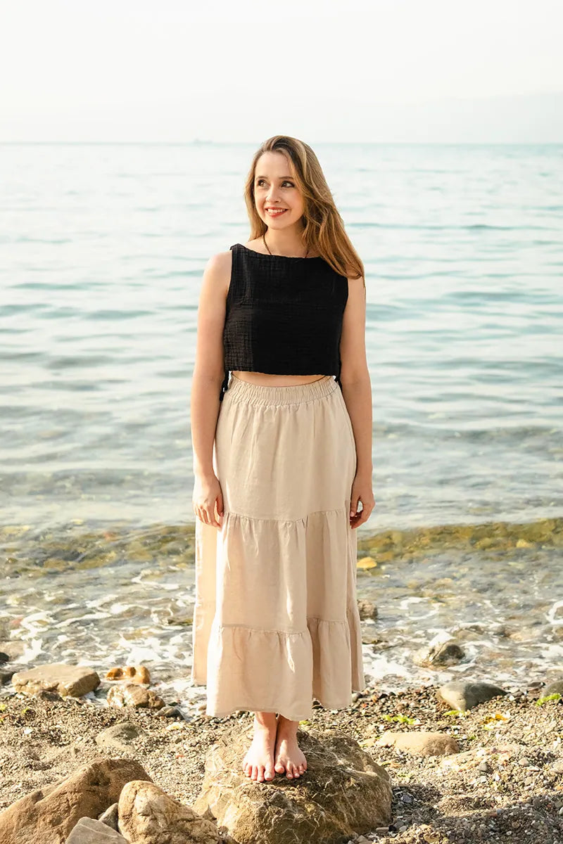 Woman standing on a rocky beach wearing a black cotton crop top and beige skirt.
