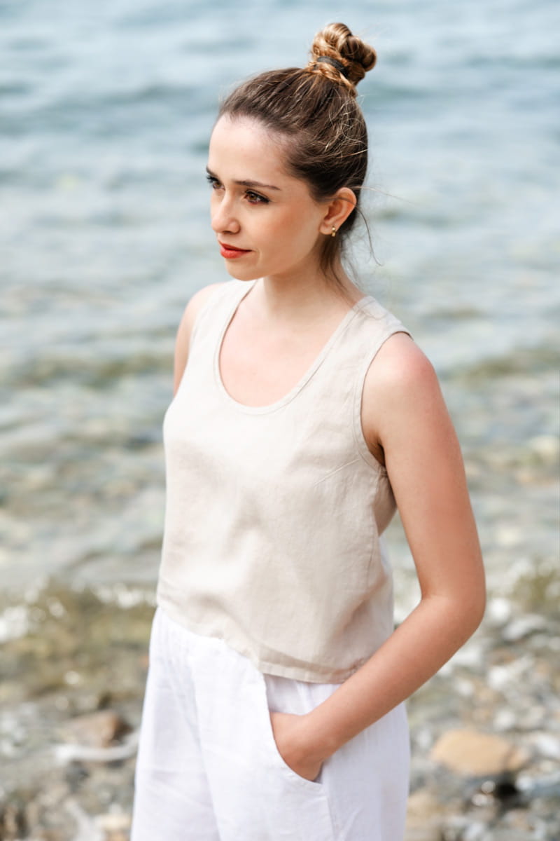 Woman wearing a beige sleeveless top and white pants standing by a body of water.