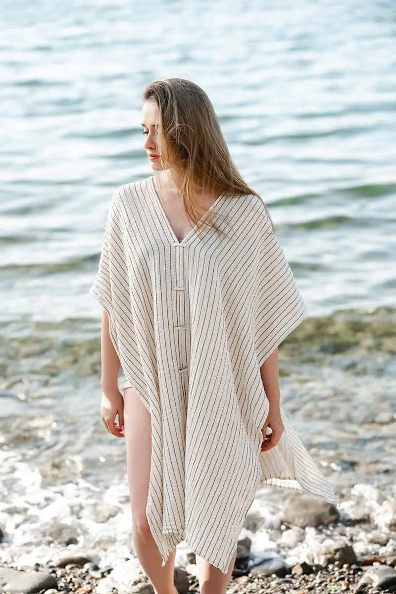 Woman wearing a beige striped poncho standing on a pebbly beach with water in the background
