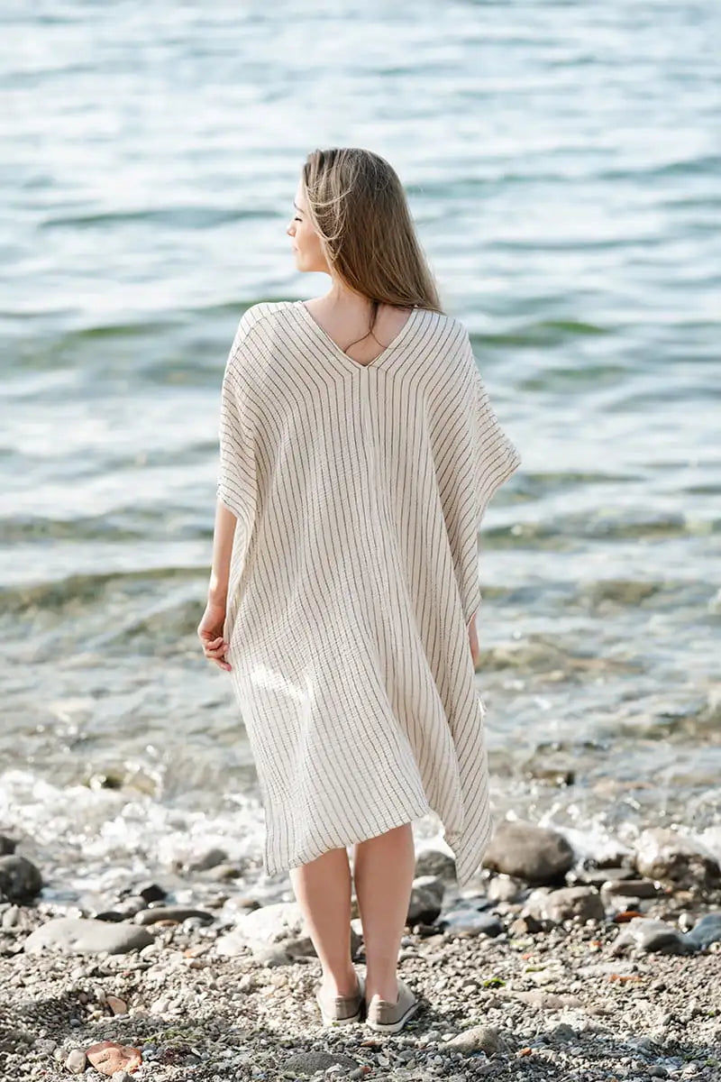Woman wearing a striped swimsuit cover up standing on a rocky beach with water in the background