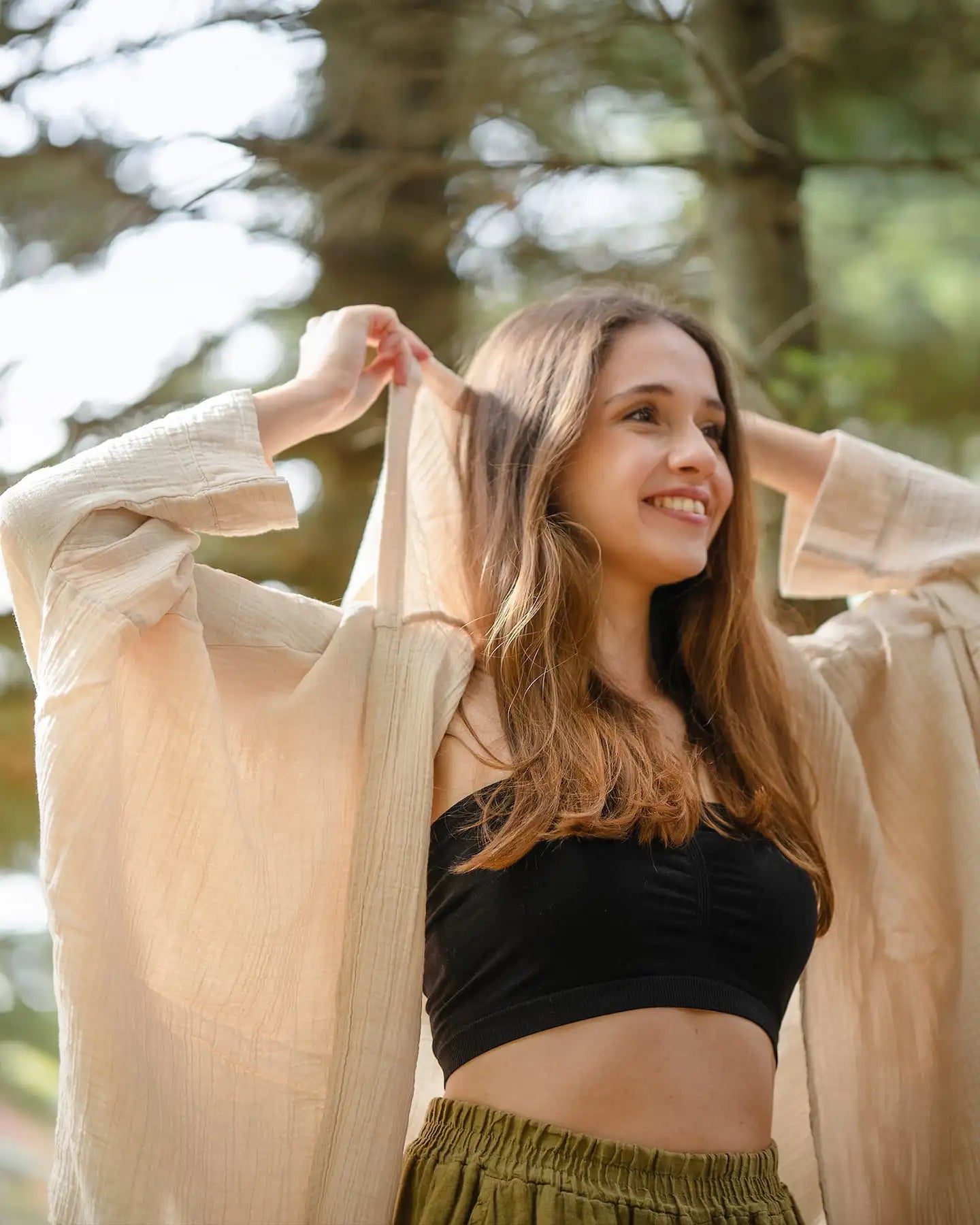 Woman holding a beige cardigan outdoors with blurred trees in the background