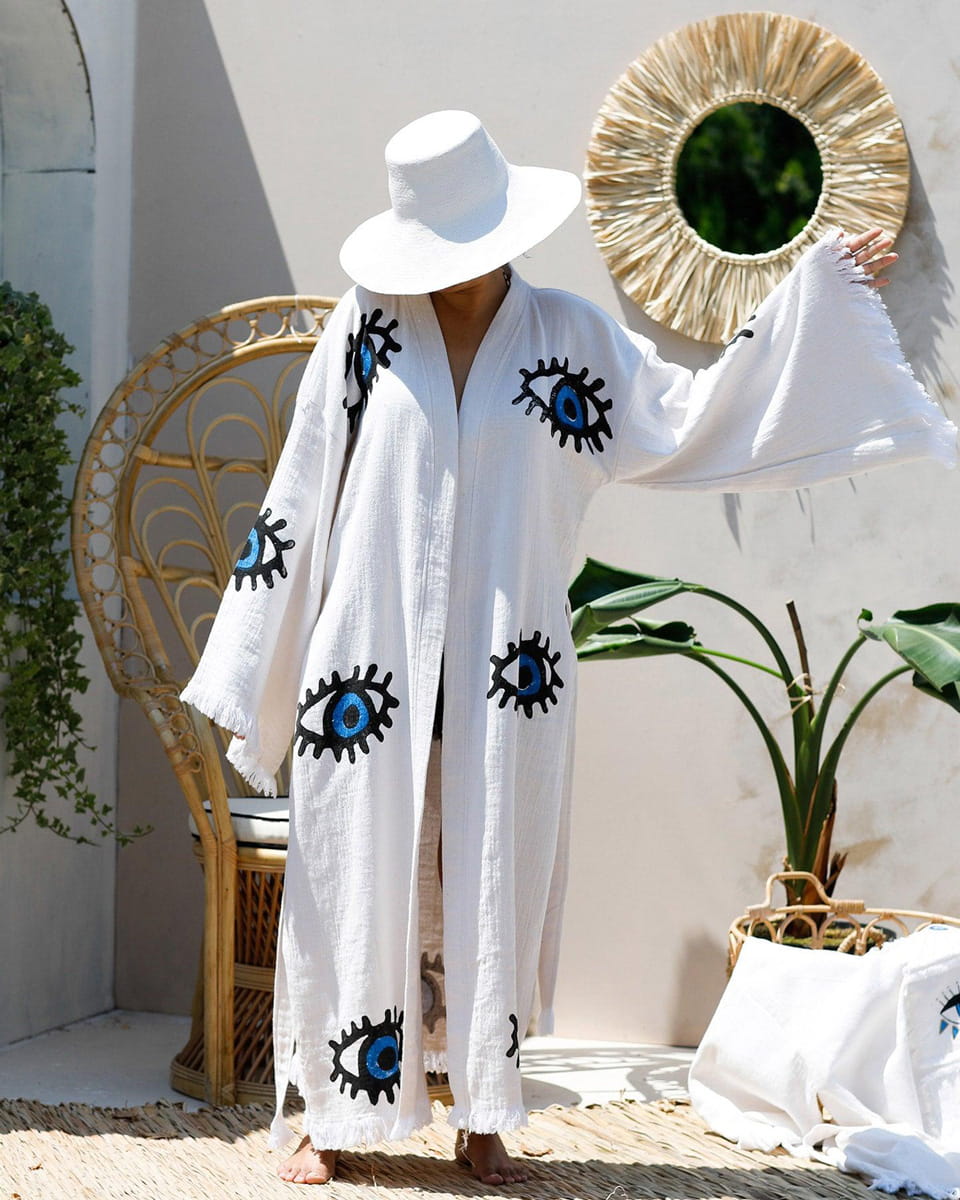 Person wearing a white outfit with blue eye designs, standing in a decorative outdoor setting.