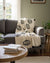 Gray sofa with a decorative blanket featuring Evil eye patterns and pom poms in a living room setting.