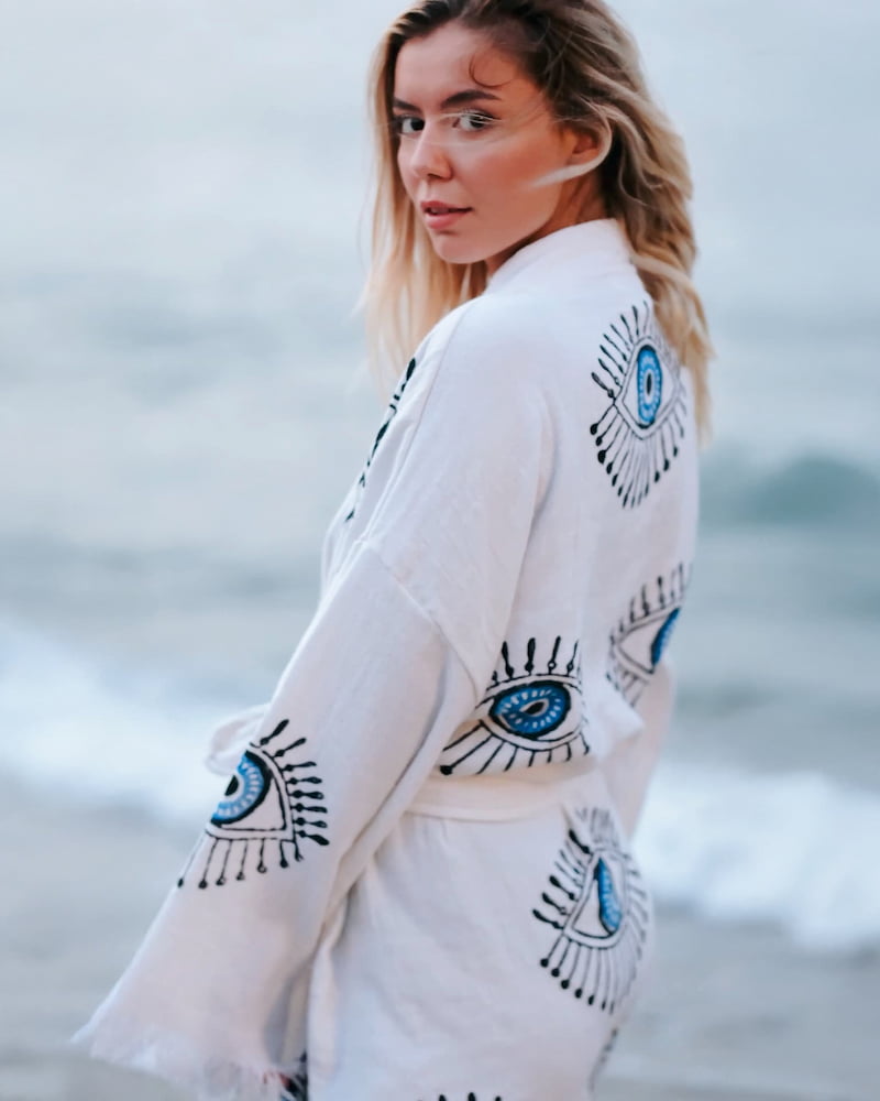 Woman wearing a white beach cover up with blue evil eye pattern against a blurred natural background