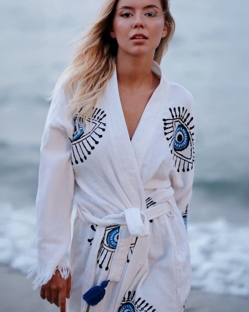 Woman wearing a white terry robe with blue and black evil eye designs on a beach.