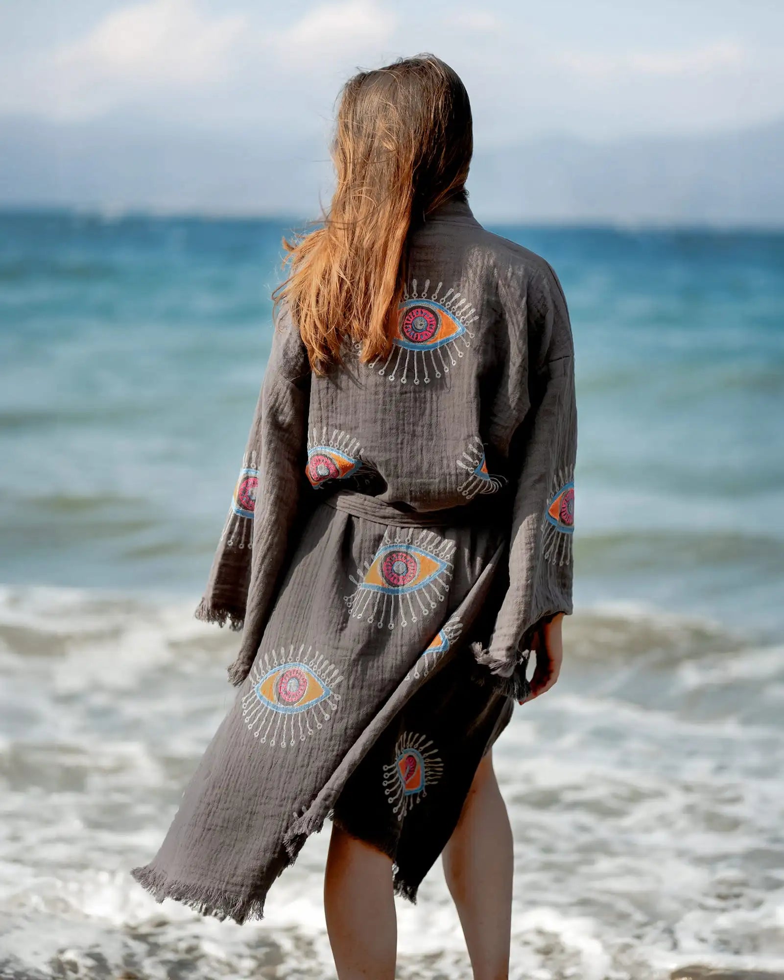 Person wearing a dark gray evil eye kimono with colorful eye designs on a beach.