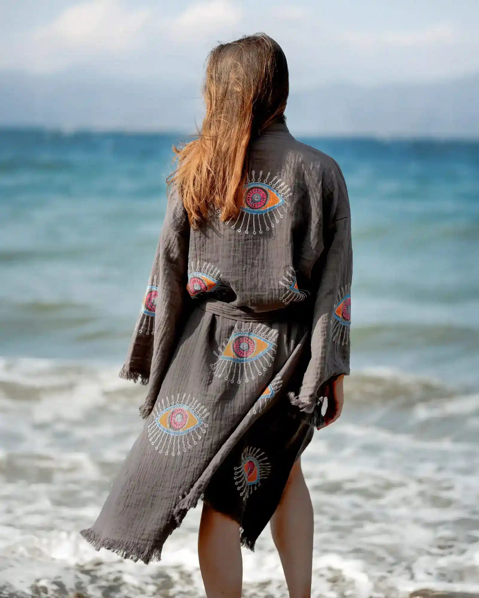 Person wearing a dark gray evil eye kimono with colorful eye designs on a beach.