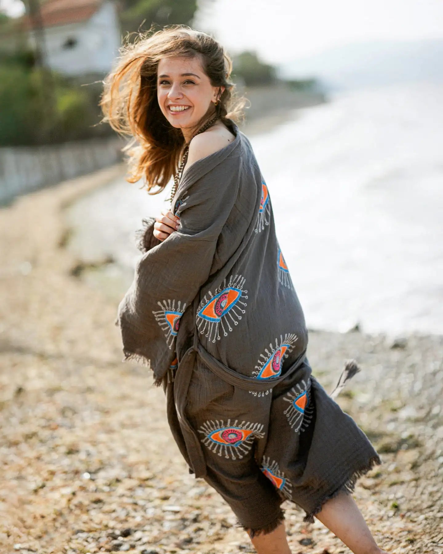 Woman wearing a patterned grey evil eye kimono poncho by a body of water