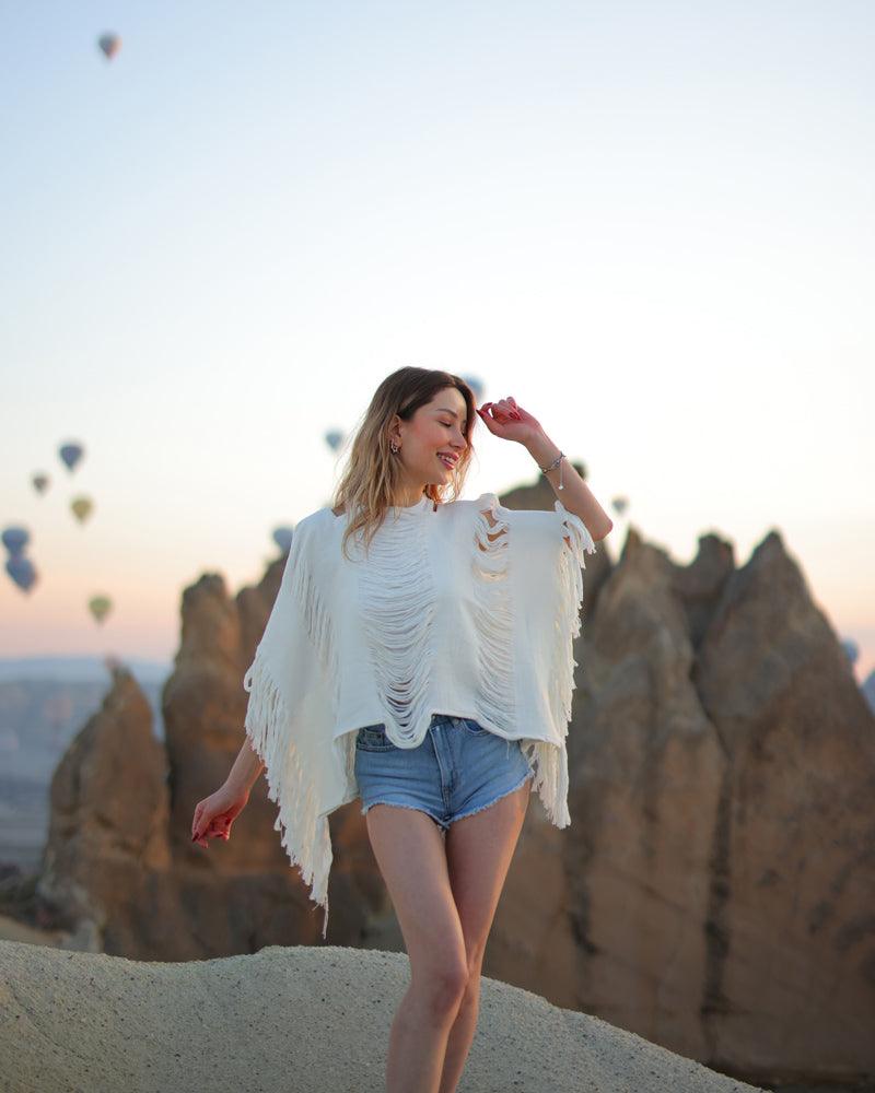 A person standing in front of a scenic backdrop featuring hot air balloons and rock formations wearing a white frayed robe with long sleeves and shorts.