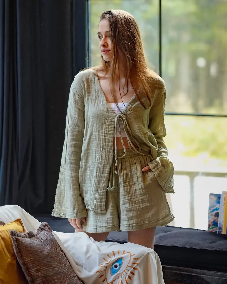 Woman in a green muslin loungewear outfit standing by a window with a blurred outdoor view.
