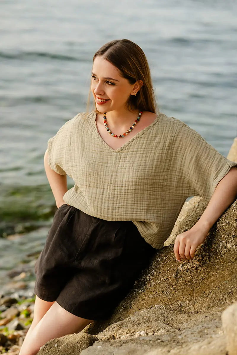 Woman sitting on a rock by the water wearing a green double gauze top and black shorts.