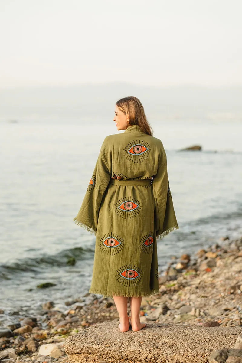 Woman wearing a green robe with eye patterns standing on a rocky beach.