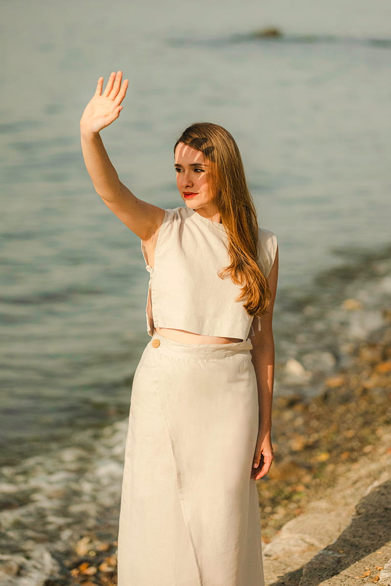 Woman in a linen crop tank top standing by a body of water, waving.