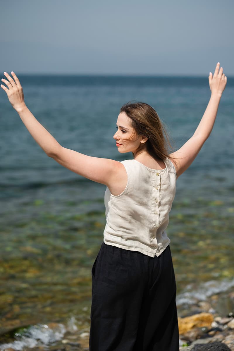 A woman in a sleeveless linen top with back buttons raises her arms by the sea
