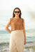 Woman wearing sunglasses and a brown linen top with beige pants standing on a beach.
