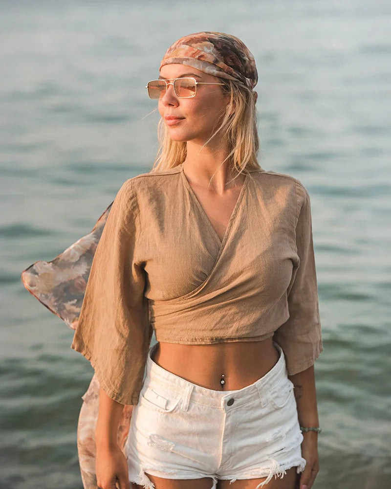Woman standing by a body of water wearing a brown tie back linen crop top and white shorts.