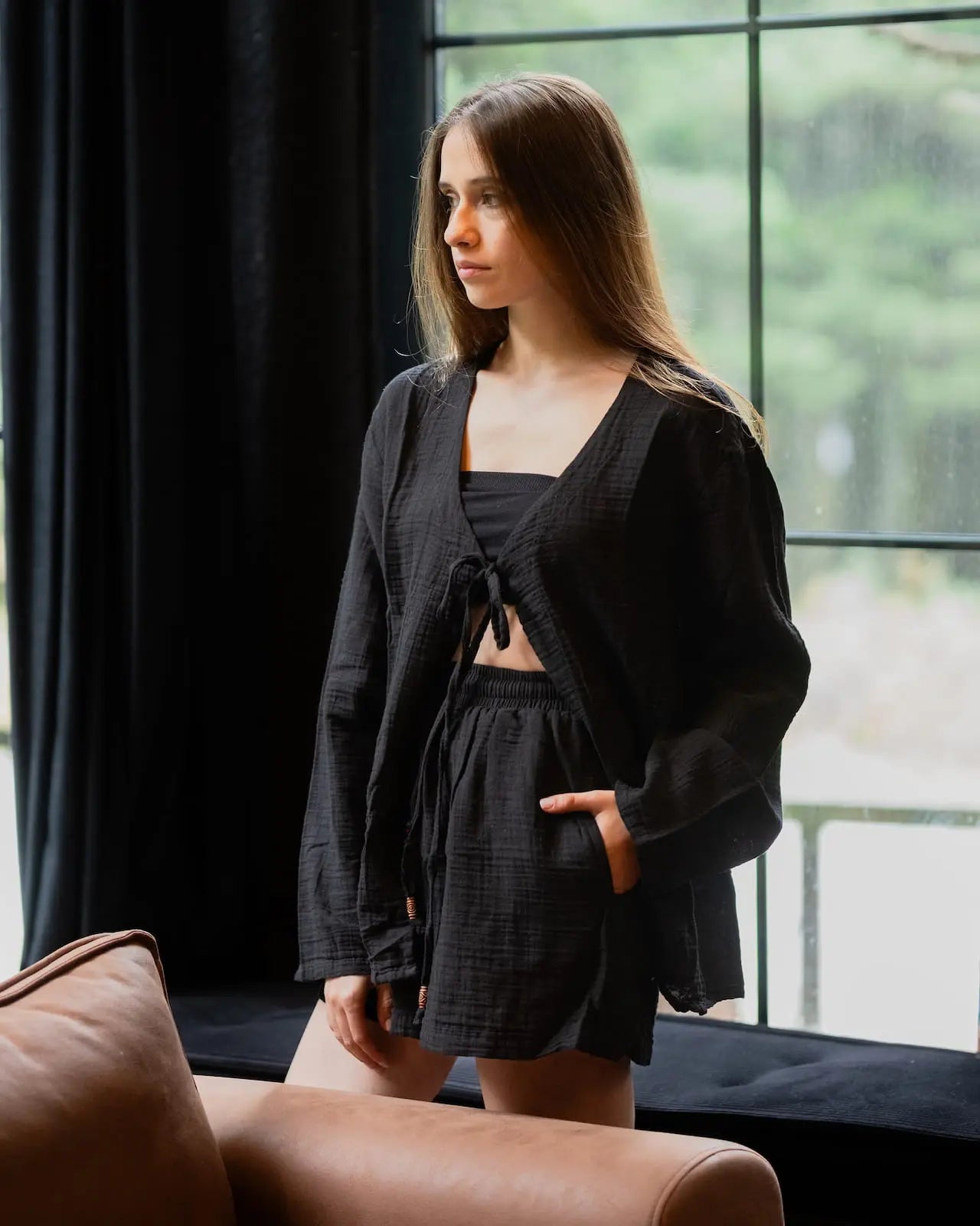 Woman in luxury black Loungewear Set standing in a room with large windows and a brown couch.