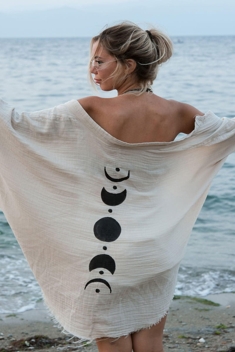 Woman standing on a beach wearing a beige cover-up with black moon phases design.