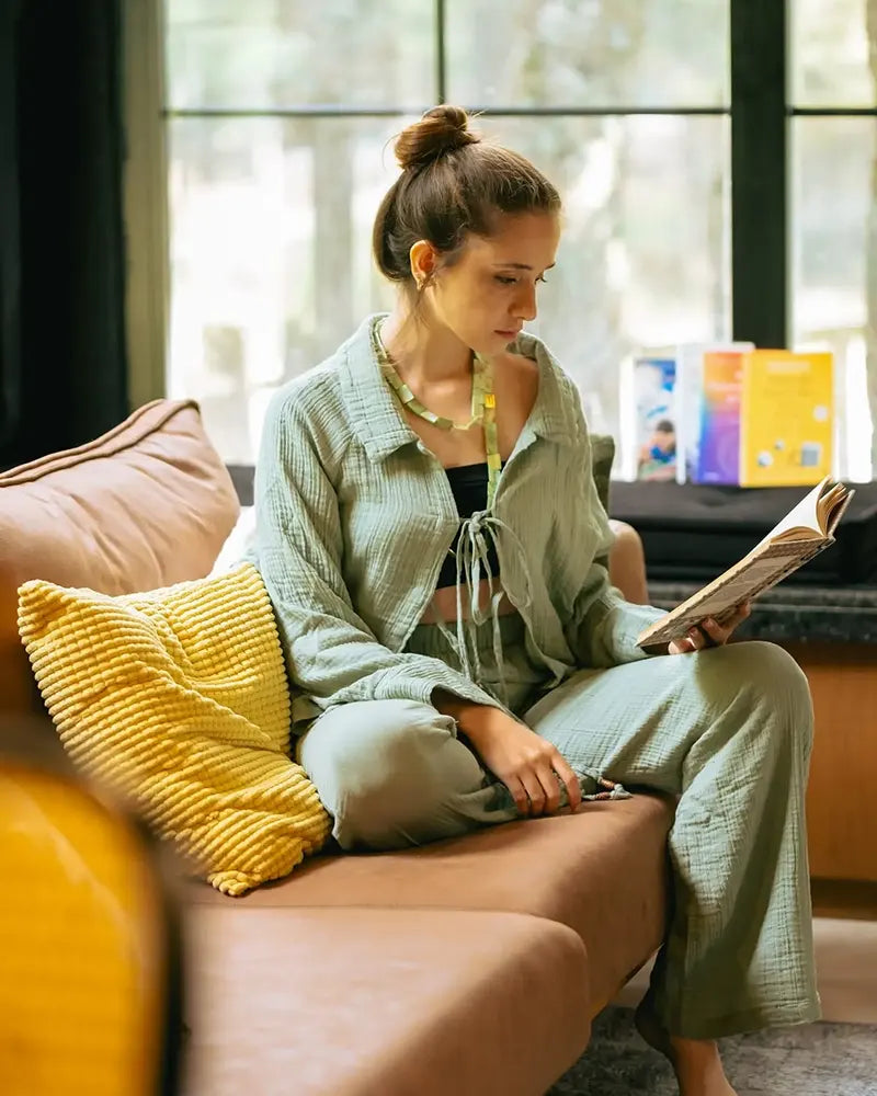 A woman dressed in muslin cotton loungewear is seated on a couch with yellow cushions, reading a book