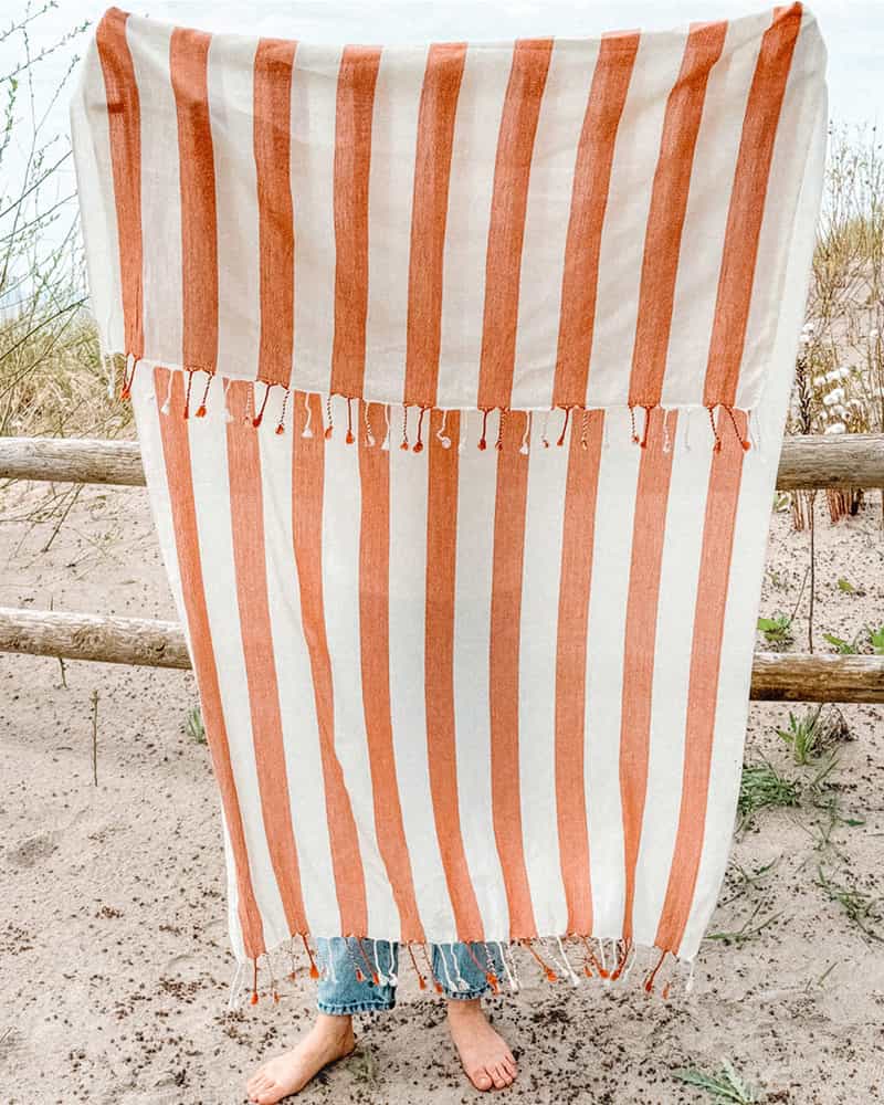 Person standing on a sandy beach with an orange and white striped towel draped over them.