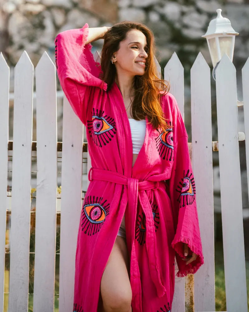 Woman wearing a bright pink robe with eye designs in front of a white picket fence.