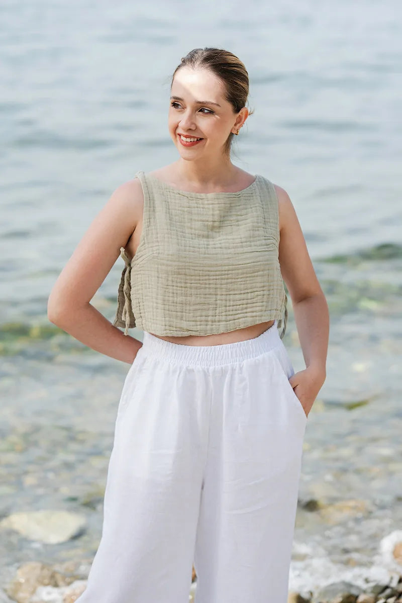 Woman wearing a green double gauze top and white pants standing by a body of water.