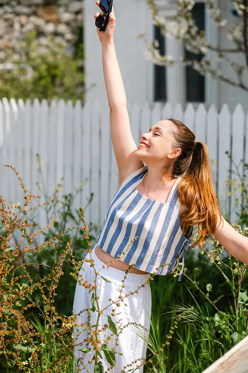 Woman in a striped blue and white top and white pants standing outdoors with a white fence and greenery in the background.