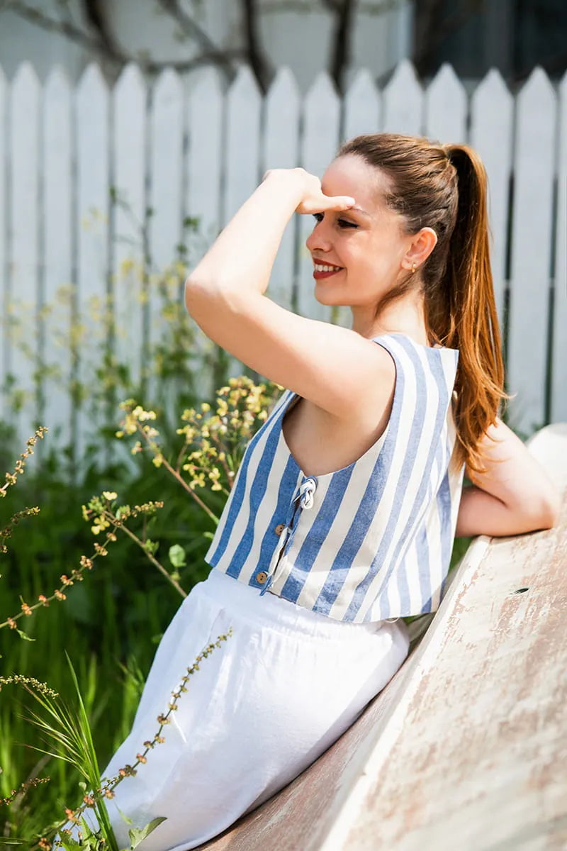 A woman is sitting on a wooden bench. She wears a blue and white striped sleeveless crop top and white pants. Behind her is a white picket fence and some green plants.