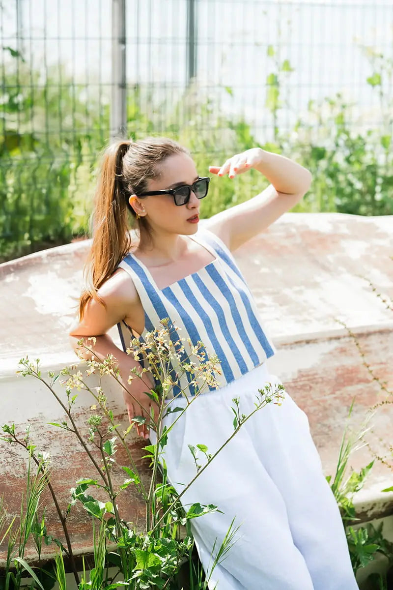 Woman in a blue and white striped crop top sitting outdoors with greenery around.