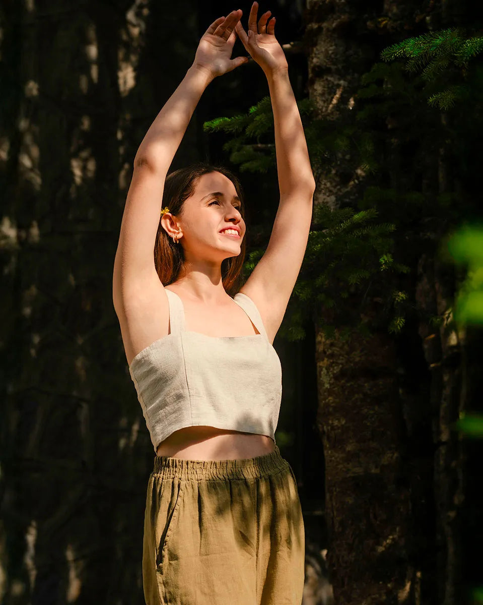 Woman in a beige crop top and green pants standing with arms raised in a forest setting