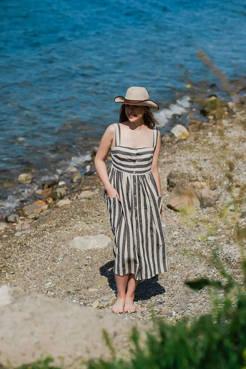 Woman in a striped dress and hat standing on a pebbly beach with water in the background