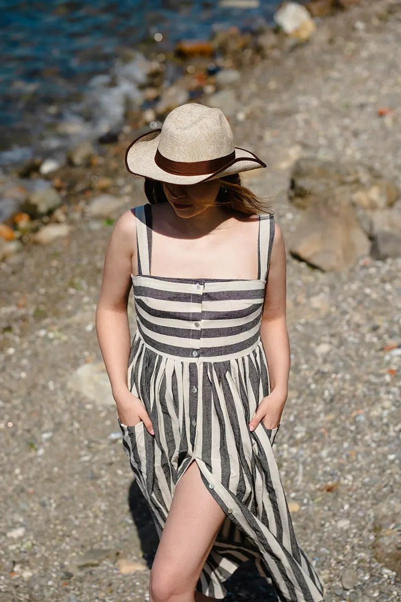 Woman in a Striped Button Down Midi Dress and hat walking on a pebbly beach.