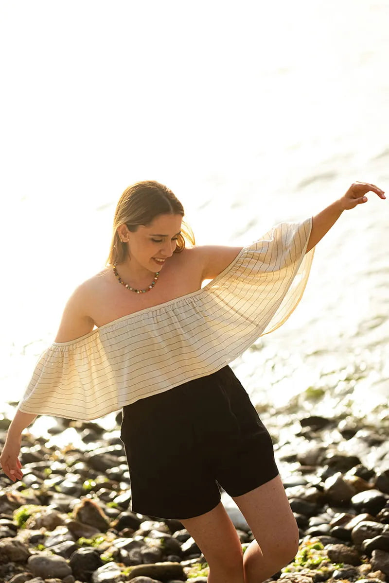 Woman wearing a striped off-shoulder top and black shorts on a rocky beach.