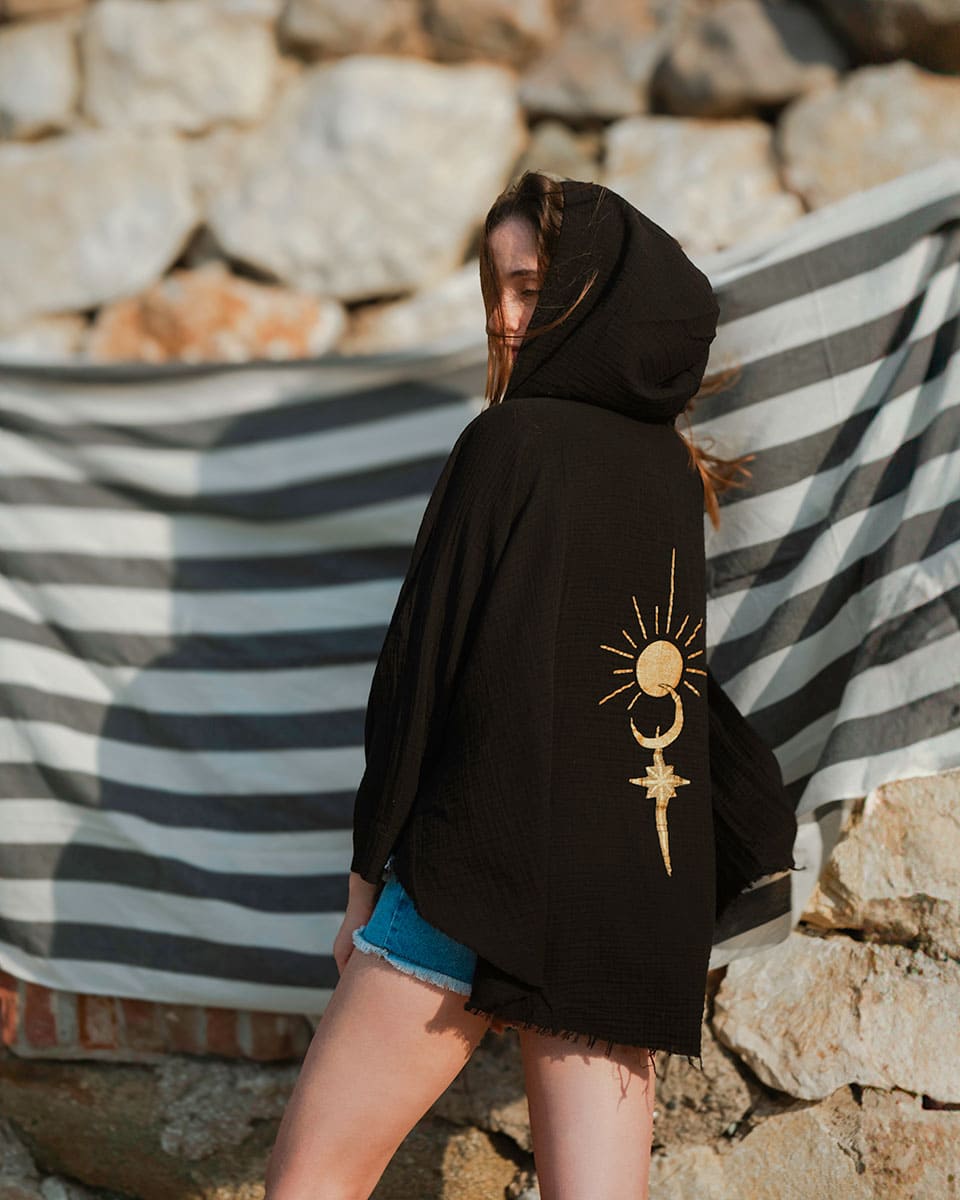 Person wearing a black hoodie with a sun and moon design, standing against a stone wall and striped fabric.