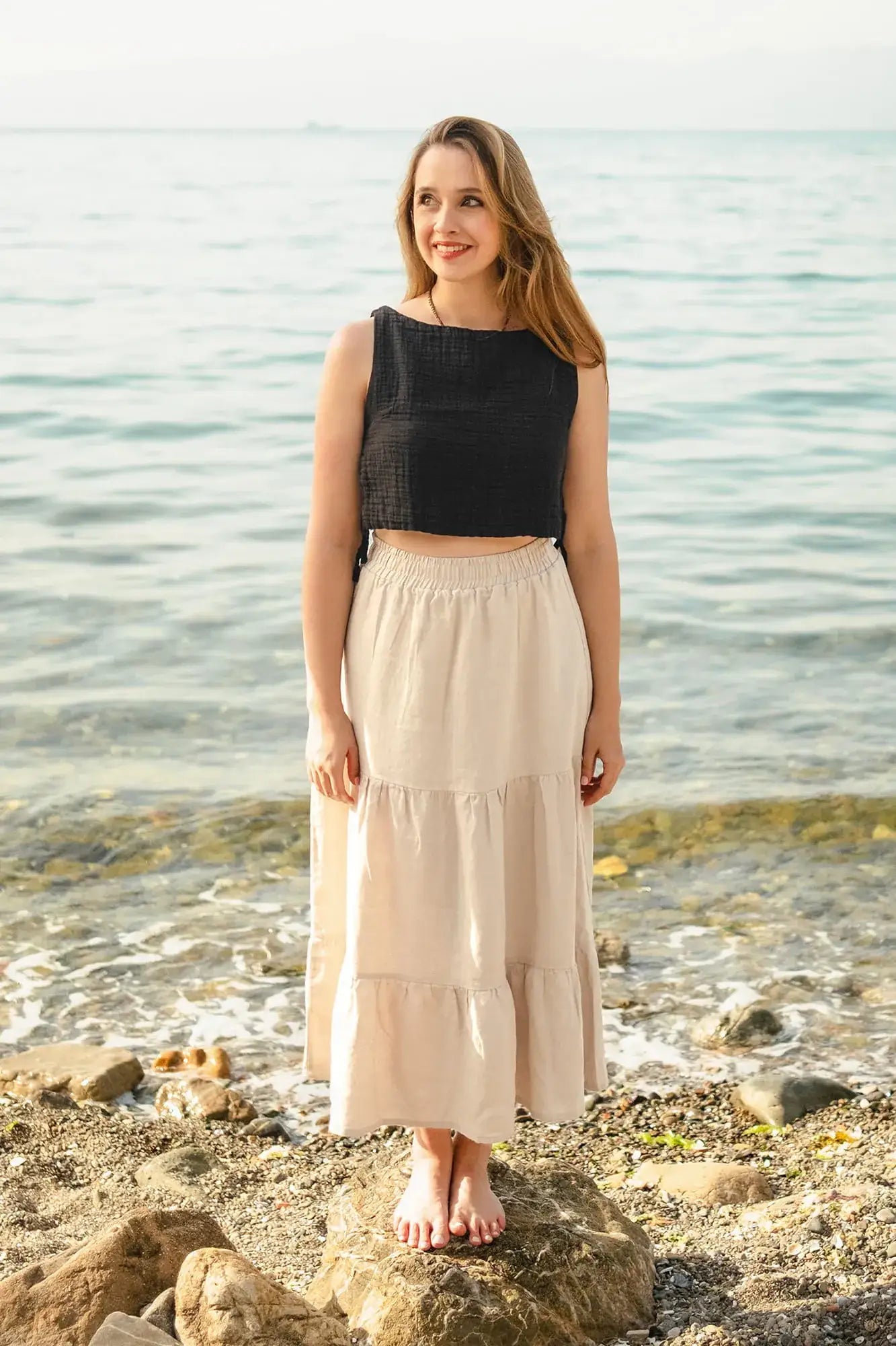 Woman standing on a rocky beach wearing a black top and beige Tiered Maxi Skirt.