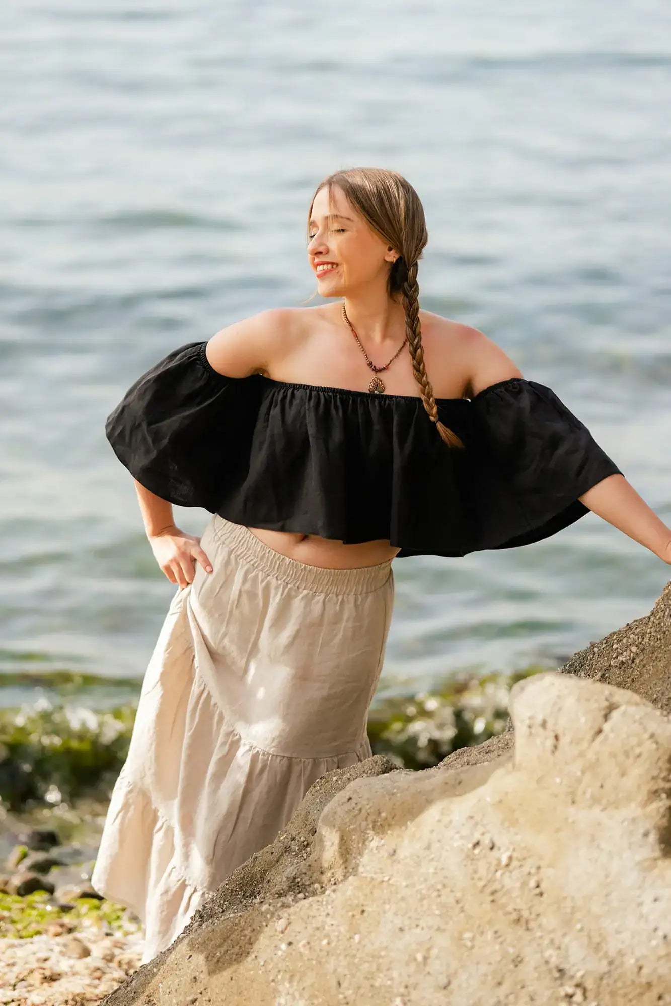 Woman in black off-shoulder top and beige maxi skirt standing on rocks by a body of water.