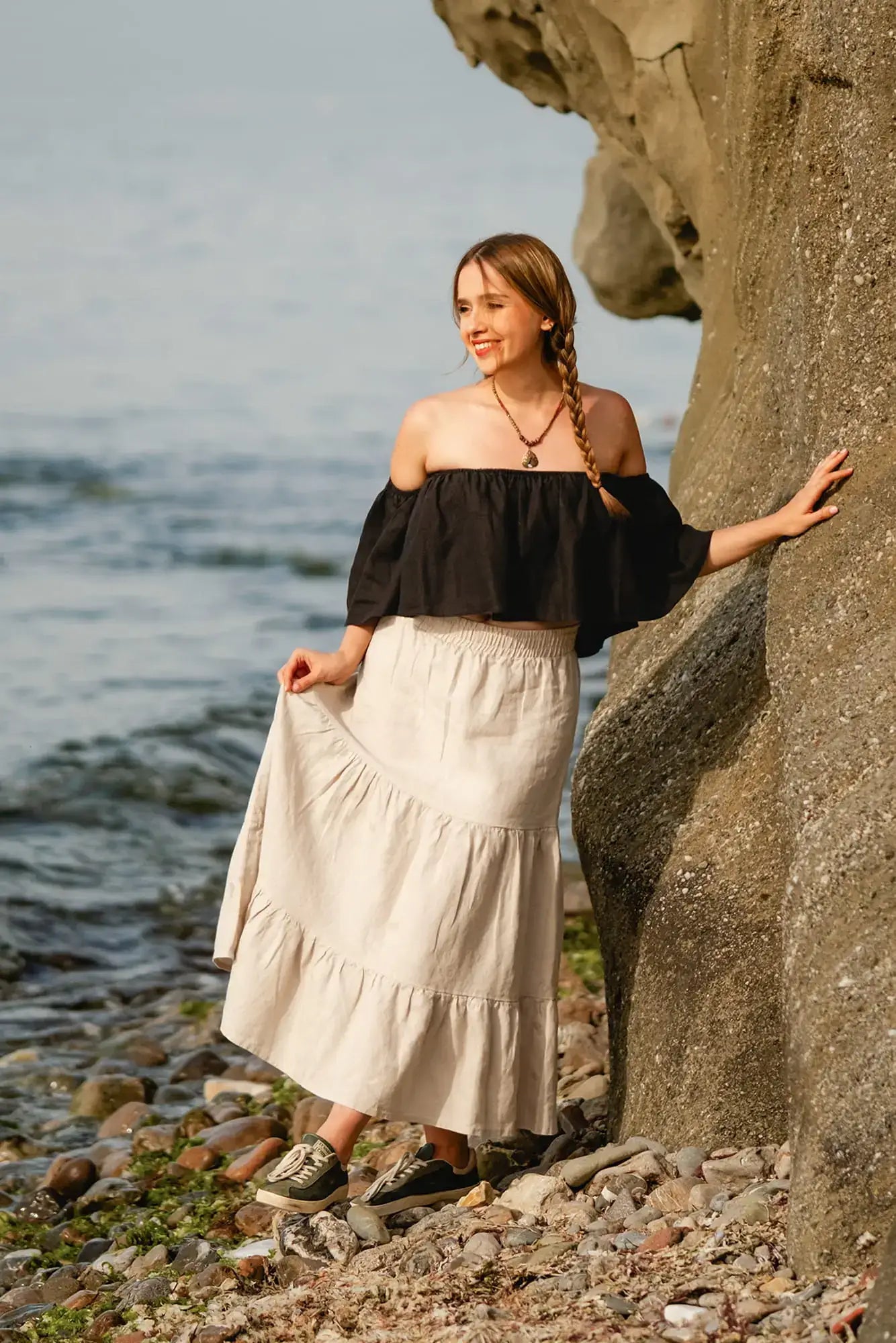 Woman in black top and Tiered Maxi skirt standing by a rocky shore with water in the background