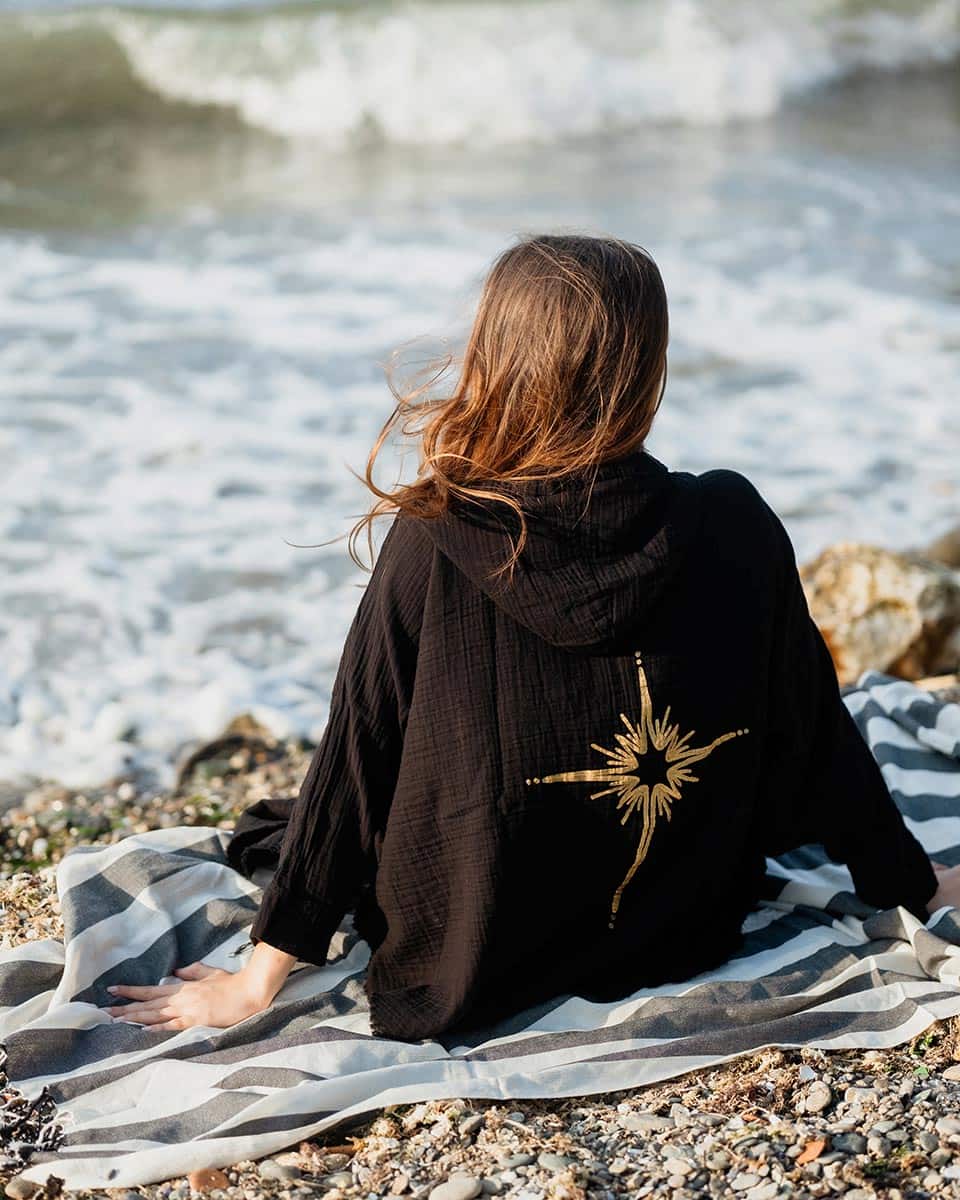 Person sitting on a beach with a black hooded garment featuring a gold star.
