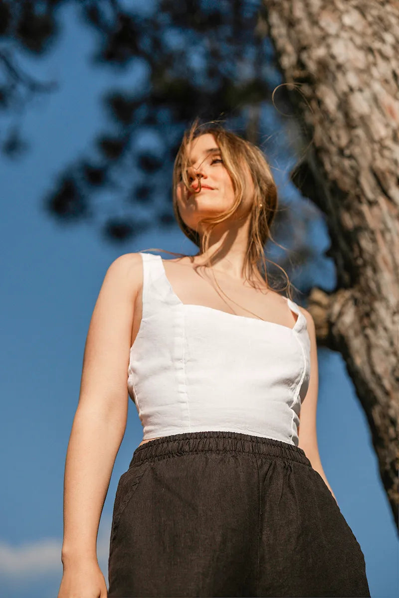 Woman wearing a white round neck crop top and black pants standing outdoors with a tree in the background