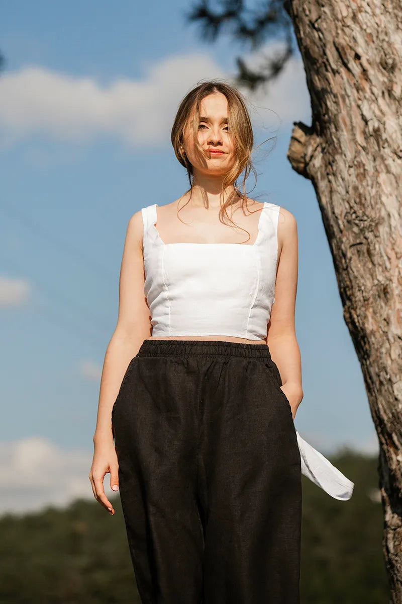 Woman in a white sleeveless crop top and black pants standing next to a tree with a blue sky background