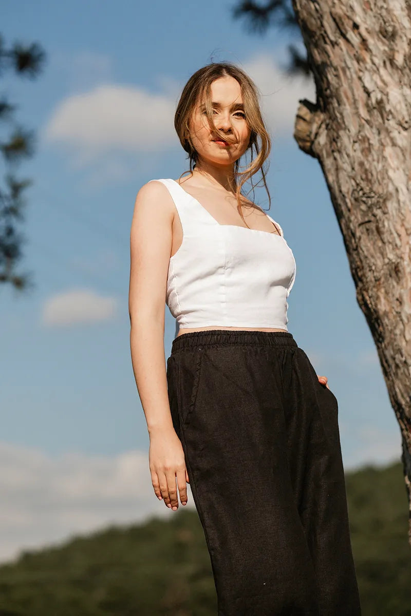 Woman in a white tie back crop top and black pants standing outdoors with trees and blue sky in the background