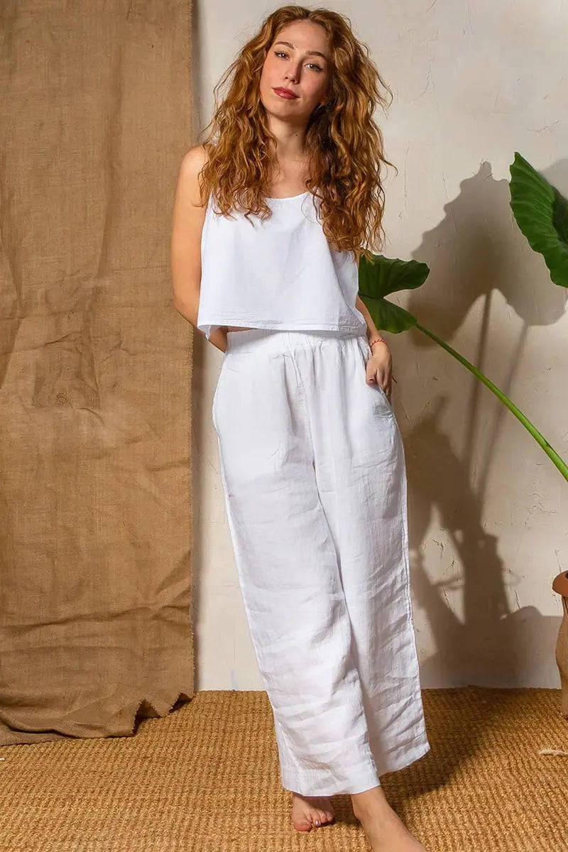 Woman wearing a white sleeveless top and White Linen Wide Leg Pants standing against a beige wall with a plant.