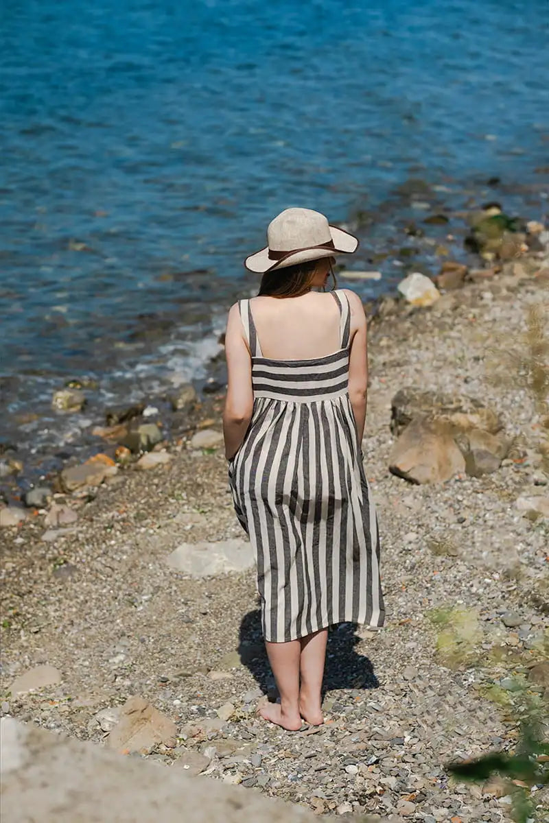 Woman in a striped linen dress and hat standing on a pebbly beach looking at the ocean.