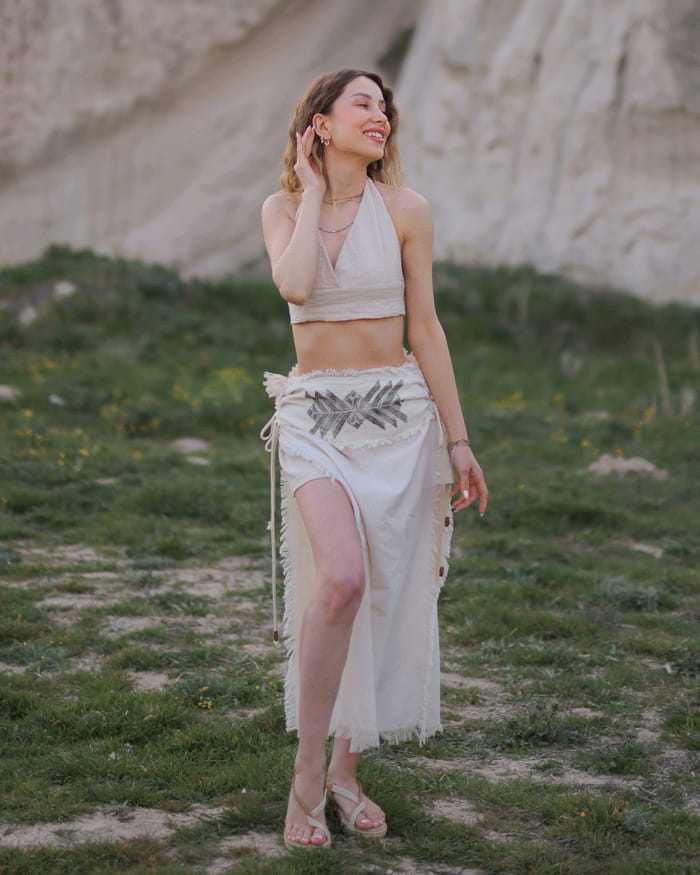 Woman in a beige top and boho skirt standing in a natural setting with green grass and rocky background.