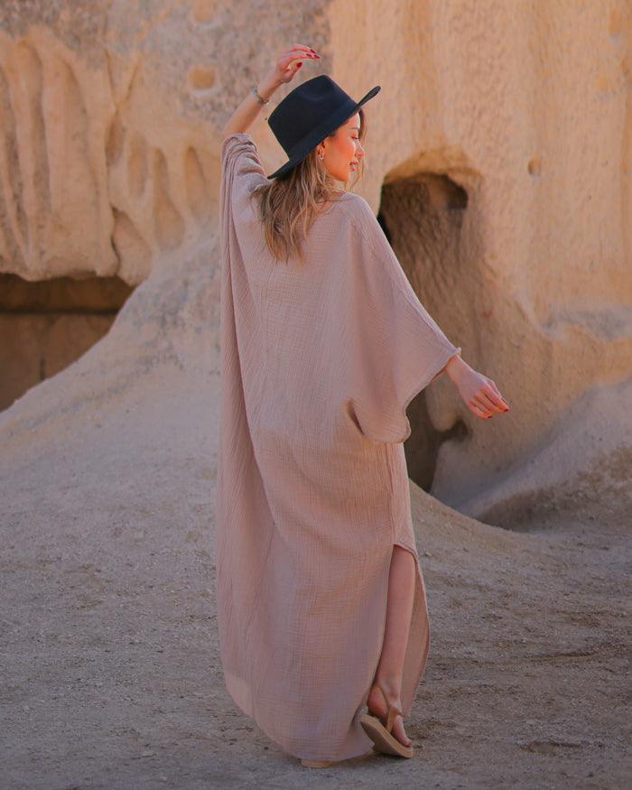 A woman in a beige maxi dress and hat strolls through a sunlit desert landscape.