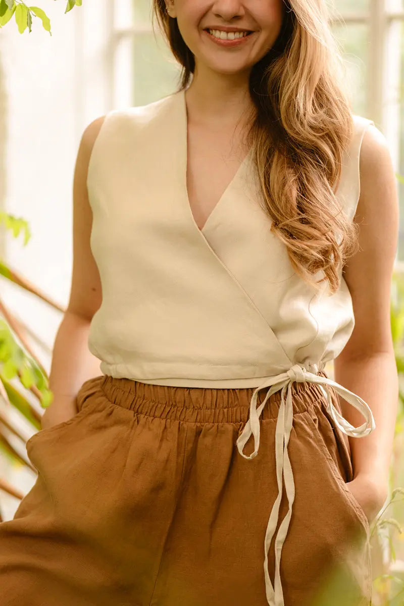 Woman wearing a beige v neck tie front sleeveless vest top and brown pants with a blurred natural background