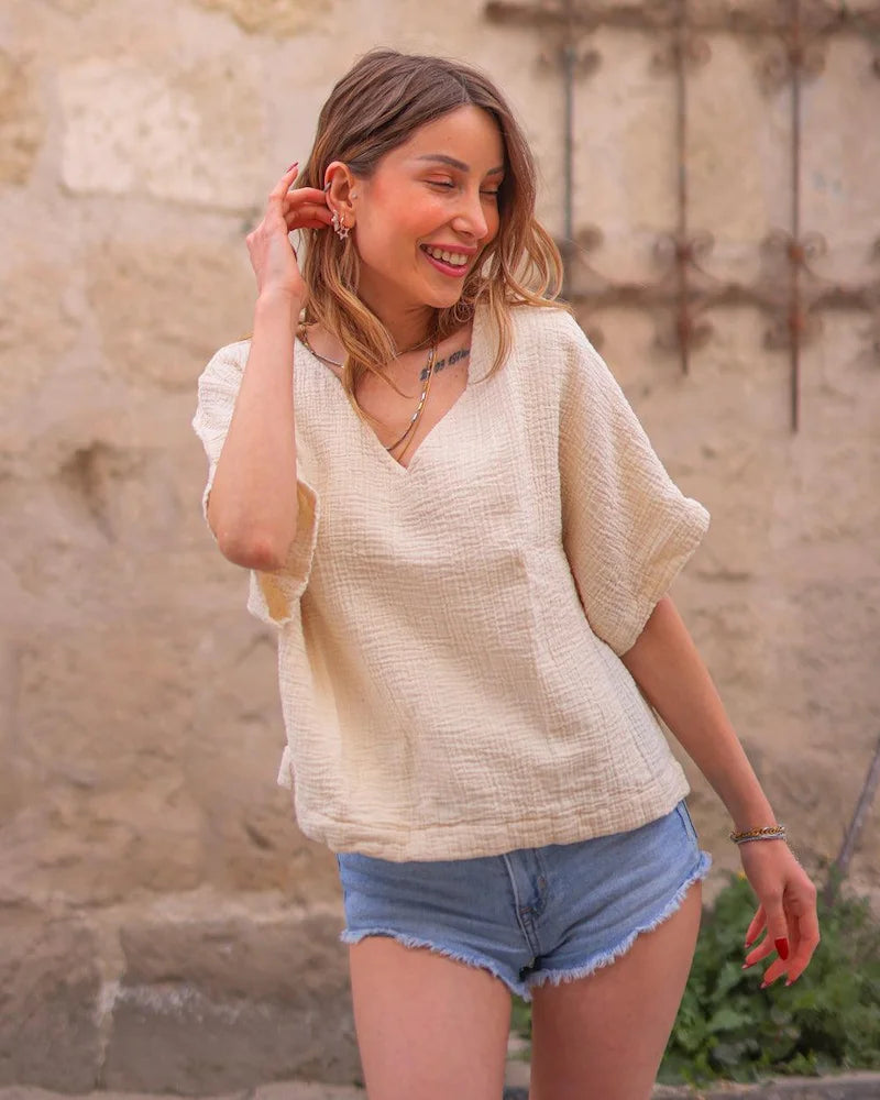 Woman wearing a beige crinkle cotton tshirt and denim shorts standing in front of a stone wall.