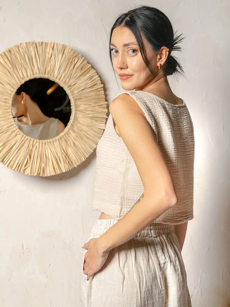 Woman in a beige top and white pants standing next to a round woven mirror on a plain wall.