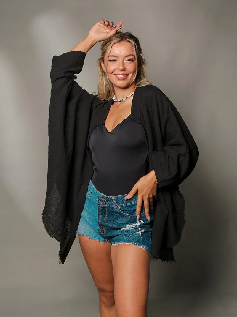 Woman wearing a black cardigan, black top, and denim shorts against a gray background