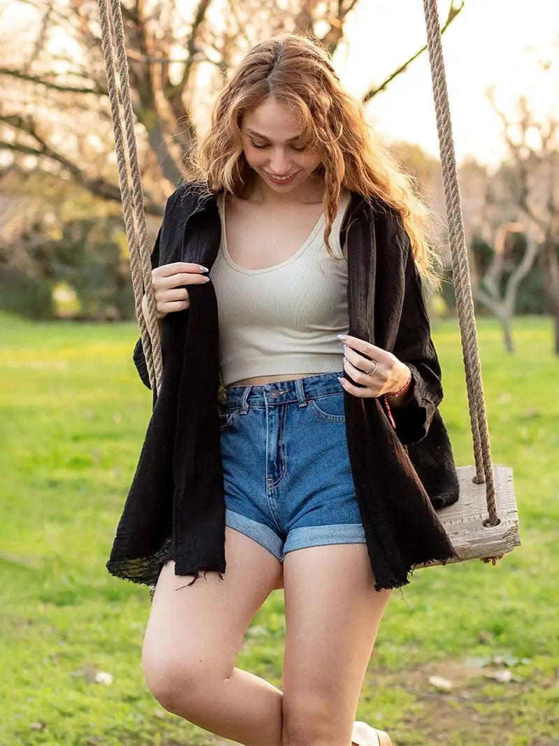 Woman on a swing in a park wearing a black cardigan, white tank top, and blue shorts.